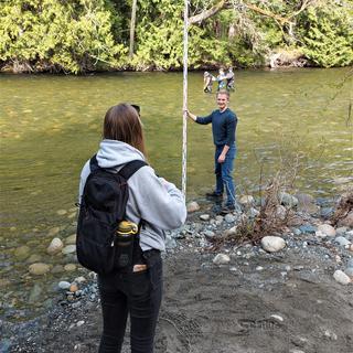 a sunny day by the river. students in the river measuring flow and depth. a student on the shore taking notes.