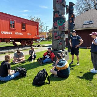 students sitting on the grass in a circle chatting on a nice sunny day in Duncan. Totem poles and a rail car are in the background