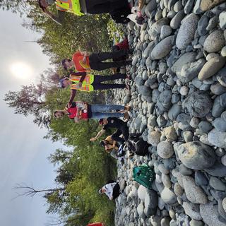 students in high visibility vests outside on a sunny day sitting and standing on cobbles and boulders with trees in the background