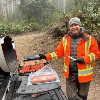 man wearing high vis cooking hot dogs on a bbq in the woods