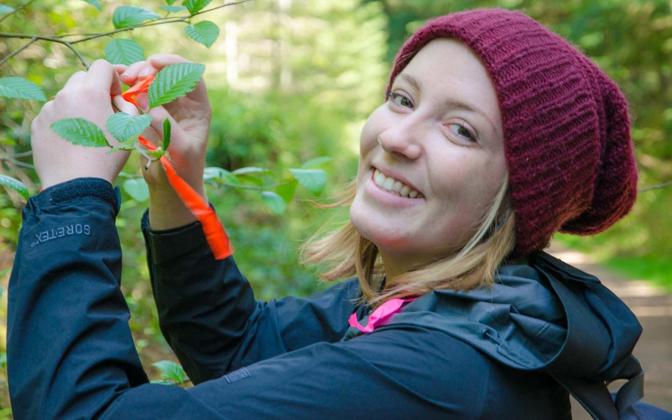 A student tying flagging tape to a branch, smiling at the camera