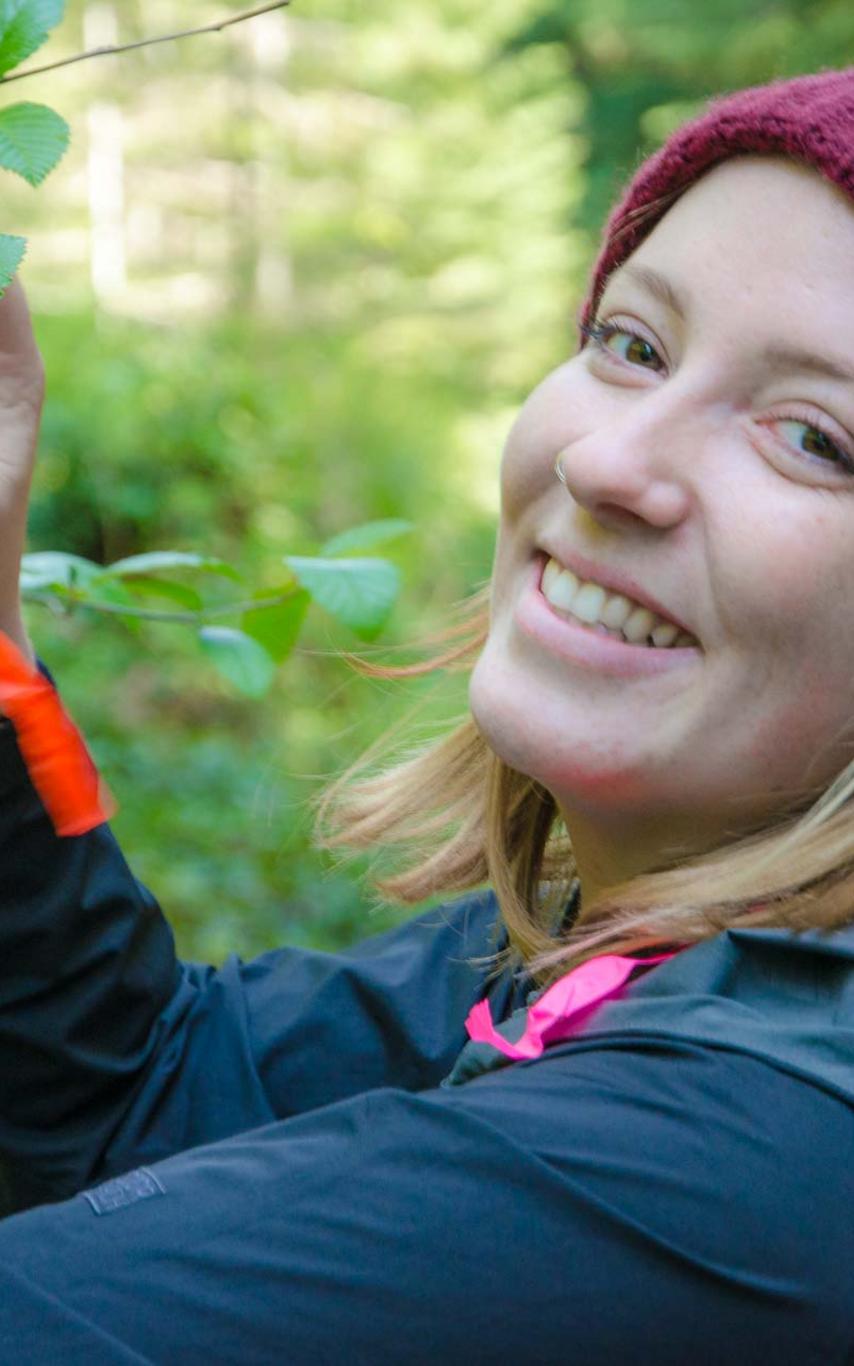 A student tying flagging tape to a branch, smiling at the camera